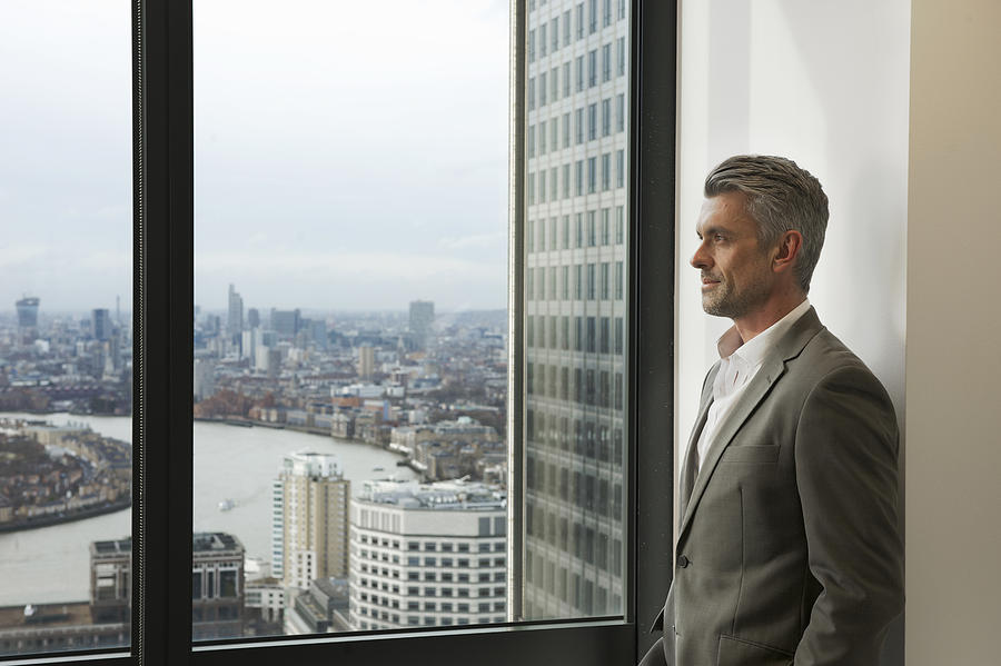 Portrait of mature businessman looking out of office window, Canary Wharf, London, UK Photograph by Andy Paul