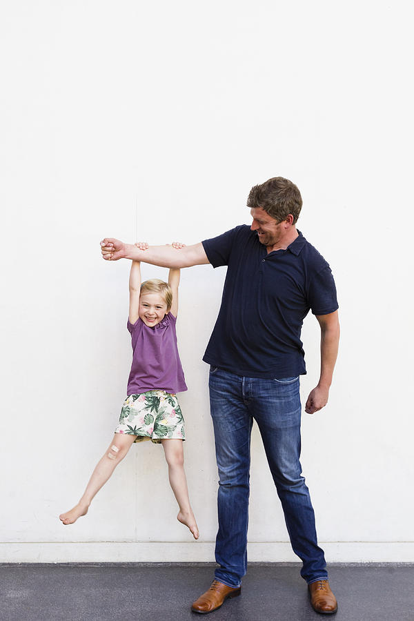 Portrait of mature man with daughter hanging from his arm in front of white wall Photograph by Emely