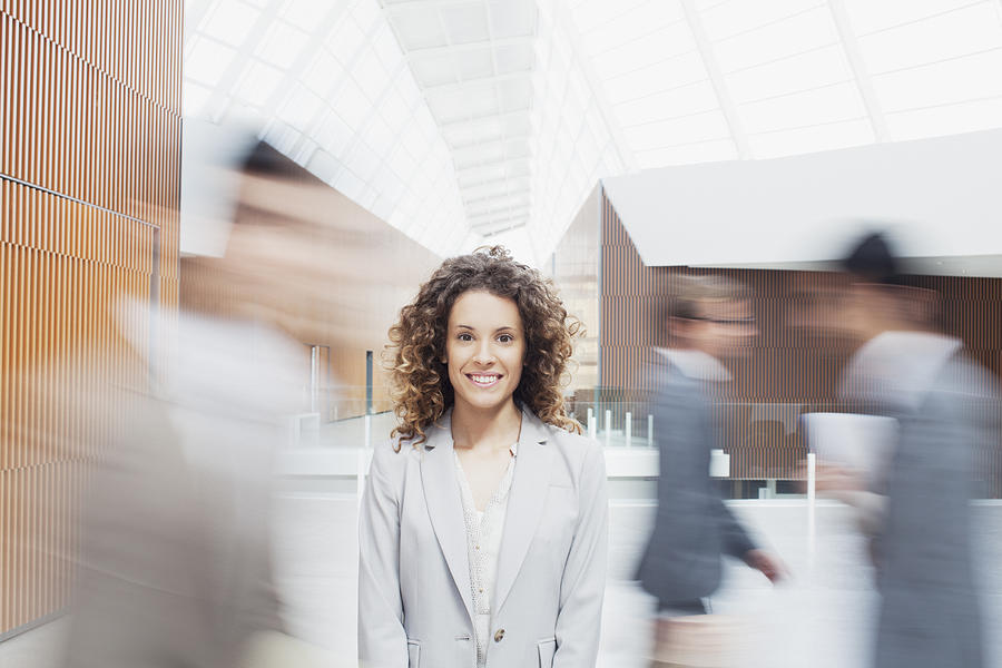 Portrait of serious woman with co-workers rushing by in lobby Photograph by Martin Barraud