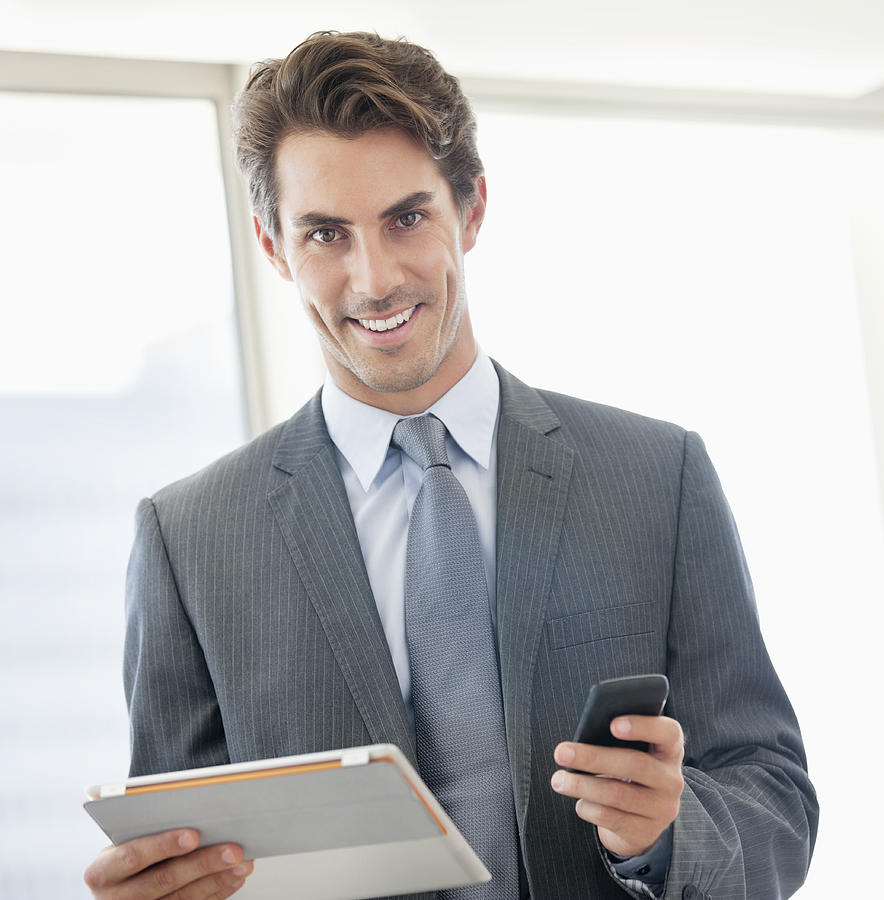 Portrait of smiling businessman holding cell phone and digital tablet Photograph by Sam Edwards