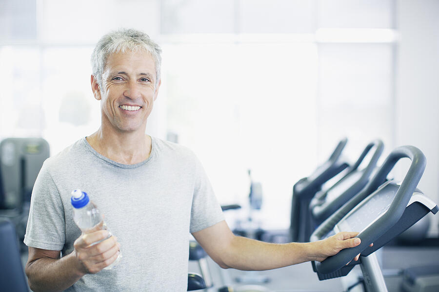 Portrait of smiling man holding water bottle on treadmill in gymnasium Photograph by Robert Daly