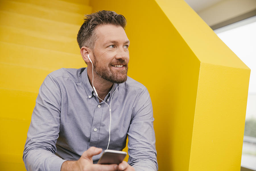 Portrait of smiling mature man hearing music with smartphone and earphones sitting on yellow stairs Photograph by Westend61