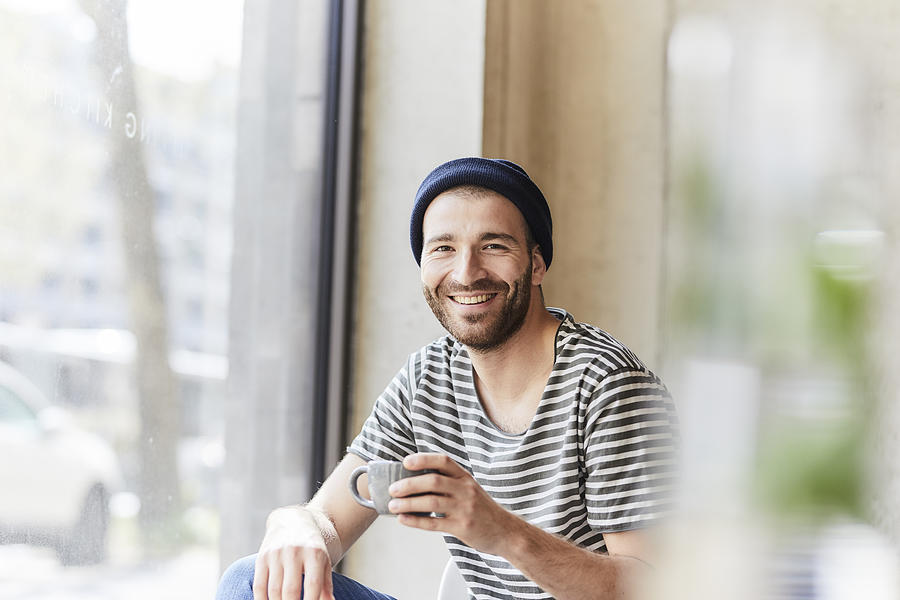 Portrait of smiling young man holding coffee cup at the window Photograph by Westend61