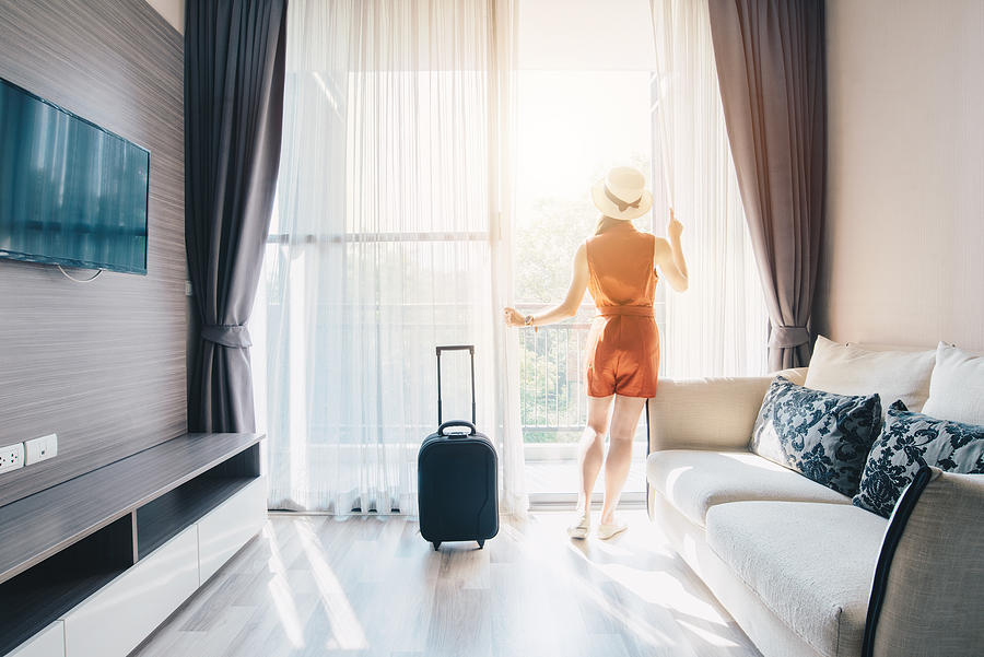 Portrait of tourist woman standing nearly window, looking to beautiful view with her luggage in hotel living room after check-in. Photograph by Boy_Anupong