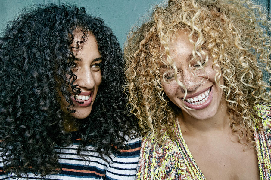 Portrait of two young women with long curly black and blond hair, smiling and laughing. Photograph by Mint Images