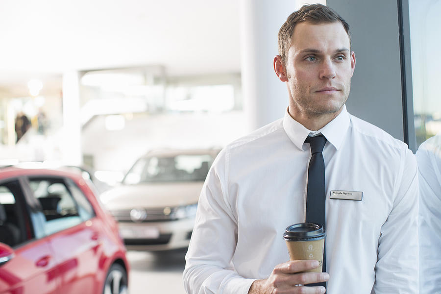 Portrait of worried salesman with takeaway coffee in car dealership Photograph by Zero Creatives