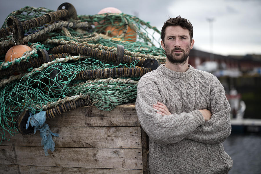 Portrait of young fisherman leaning against crate of fishing nets in harbour, Fraserburgh, Scotland Photograph by Leon Harris
