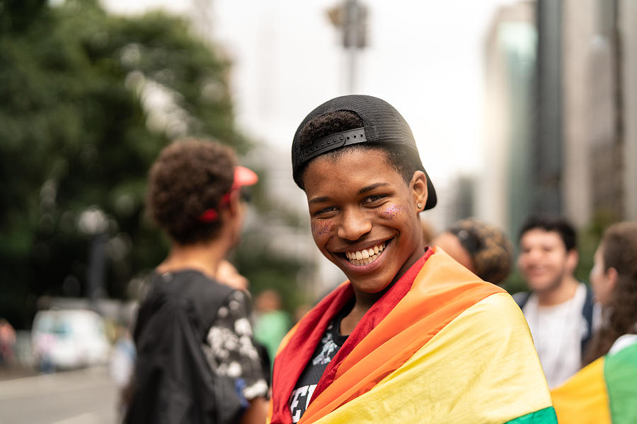 Portrait of young man with rainbow flag with friends on background at Gay Parade Photograph by FG Trade