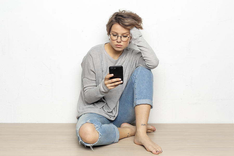 Portrait of young woman with wireless earphones sitting on the floor looking at cell phone Photograph by Westend61