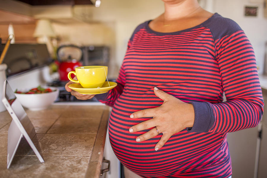Pregnant Caucasian mother holding cup of tea in kitchen Photograph by Adam Hester
