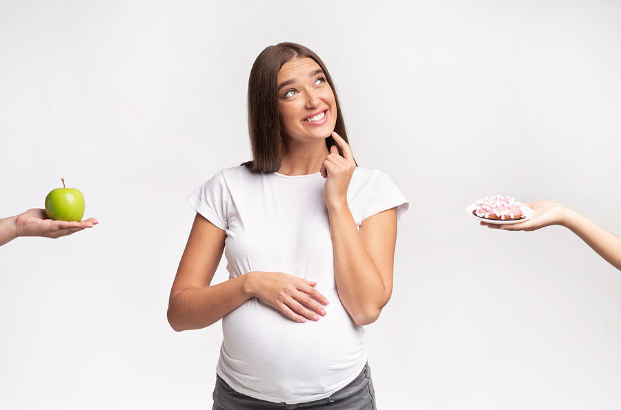 Pregnant Lady Dreaming About Sweets Choosing Doughnut Or Apple, Studio Photograph by Prostock-Studio