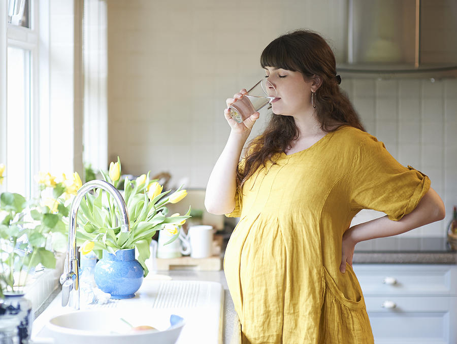 Pregnant woman drinking glass of water in kitchen at home. Photograph by Dougal Waters