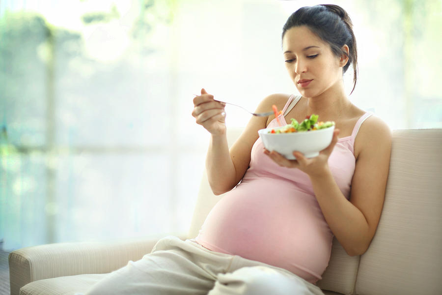 Pregnant woman relaxing at home and eating salad. Photograph by Gilaxia