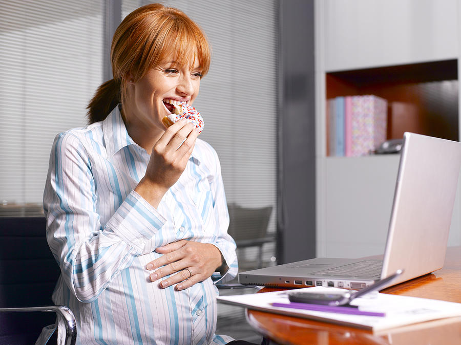 Pregnant woman sitting in front of laptop eating donut Photograph by Peter Dazeley