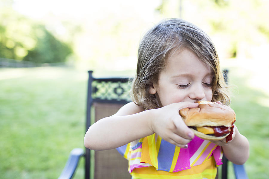 Preschool girl eating messy cheeseburger on patio Photograph by Caia Image