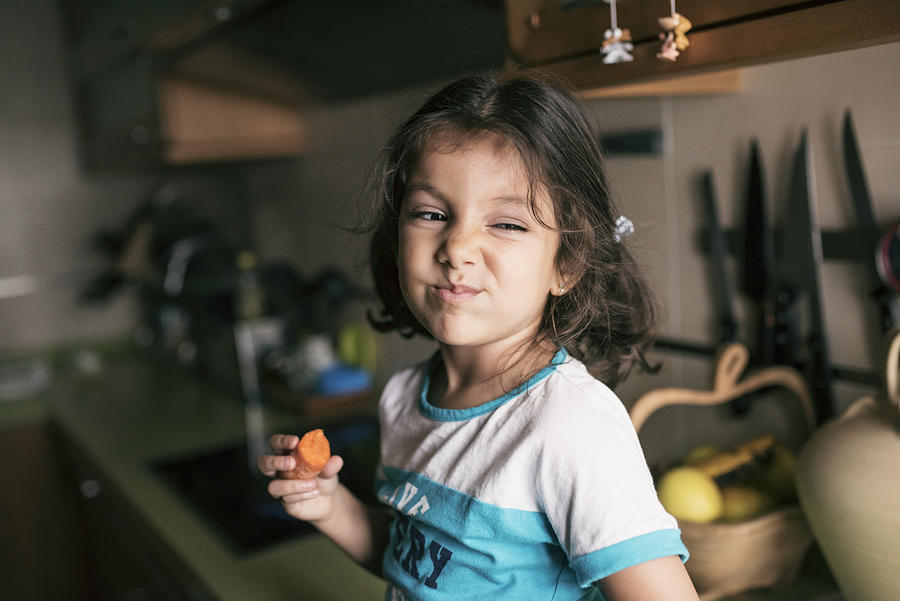 Pretty 4 years girl eating carrot in kitchen, sitting on her kitchen counter Photograph by Antonio Garcia Recena