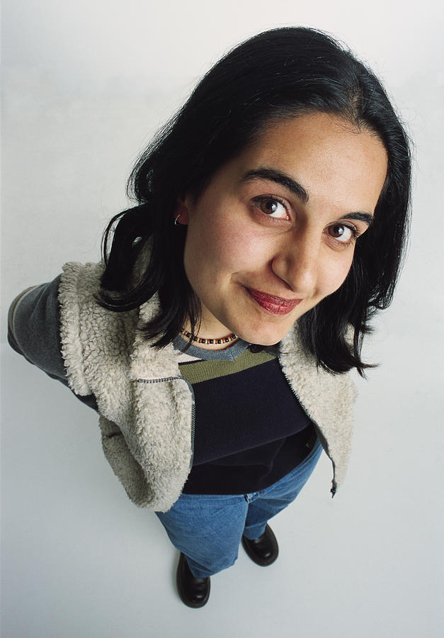 Pretty Young Adult Ethnic Female Dressed Casually In A Fleece Vest And Jeans Looks Up At The Camera With Her Head To One Side Grinning Shyly Photograph by Photodisc
