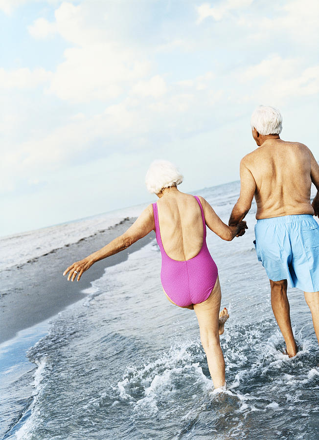 Rear View of a Senior Couple Walking in the Surf and Holding Hands Photograph by Digital Vision.