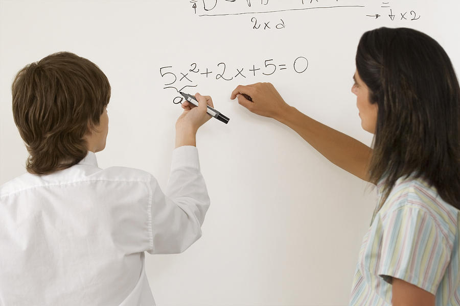 Rear view of a teenage boy solving algebra on whiteboard and a female teacher standing beside him Photograph by Glow Images
