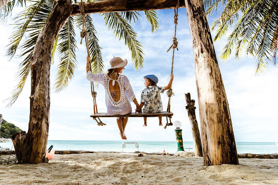 Rear view of carefree mother and son holding hands while swinging at the beach. Photograph by Skynesher