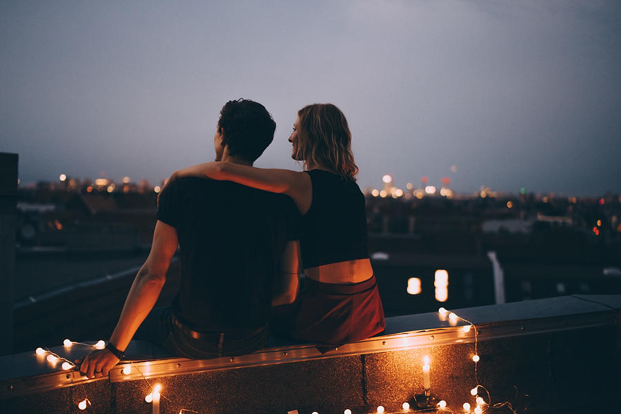 Rear view of couple with arm around sitting on illuminated terrace in city against sky Photograph by Maskot