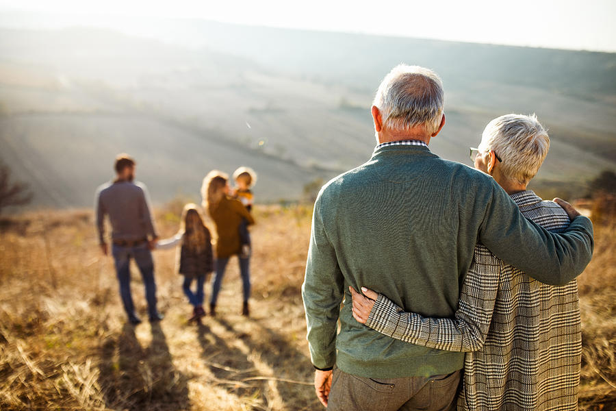 Rear view of embraced senior couple looking at their family in nature. Photograph by Skynesher