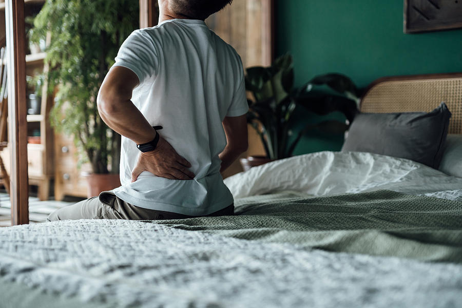 Rear view of senior Asian man suffering from backache, massaging aching muscles while sitting on bed. Elderly and health issues concept Photograph by AsiaVision