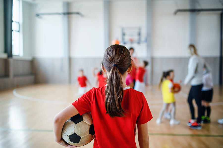 Rear view of small girl standing with ball indoors in gym class, physical education concept. Photograph by Halfpoint Images
