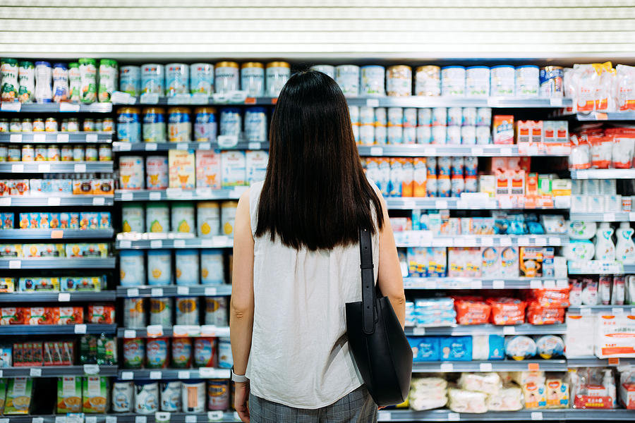 Rear view of young Asian mother groceries shopping for baby products in a supermarket. She is standing in front of the baby product aisle and have no idea which product to choose from Photograph by D3sign