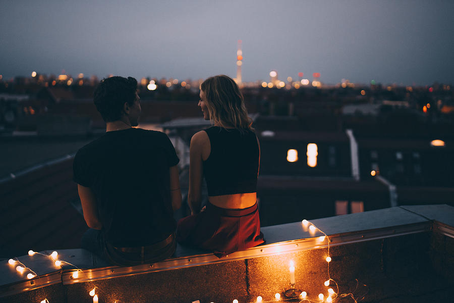 Rear view of young couple sitting on illuminated terrace in city at dusk Photograph by Maskot