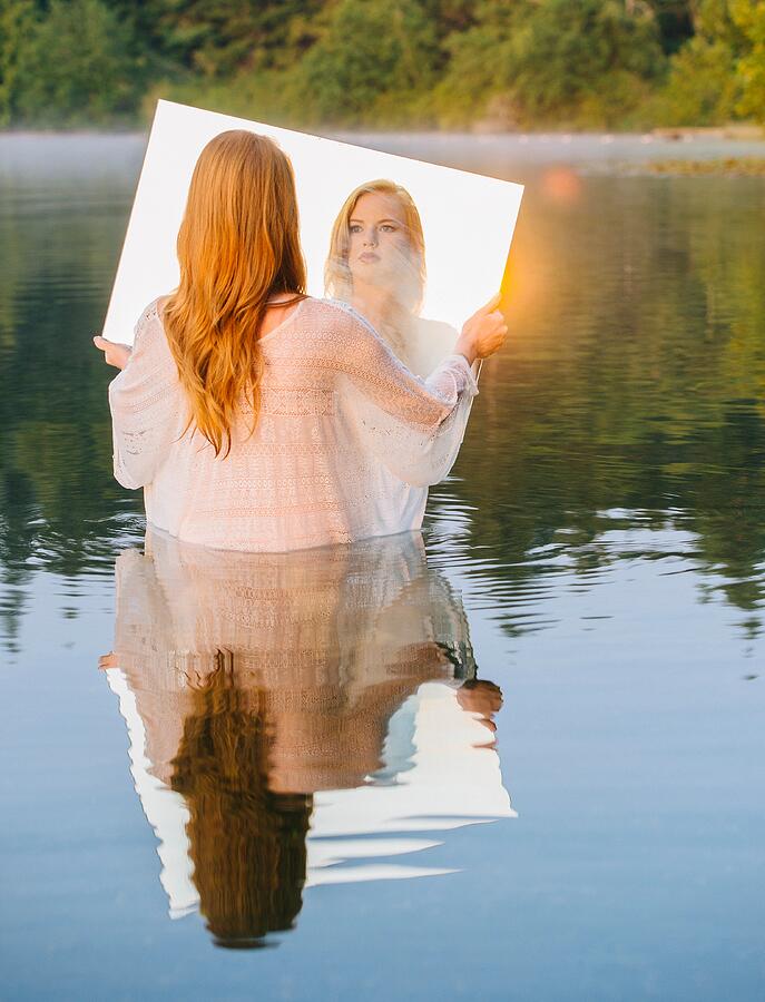 Rear view of young woman standing in lake holding mirror looking at reflection, lens flare Photograph by Pete Saloutos