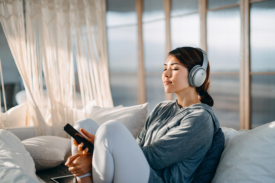 Relaxed young Asian woman with eyes closed sitting on her bed enjoying music over headphones from smartphone at home Photograph by AsiaVision