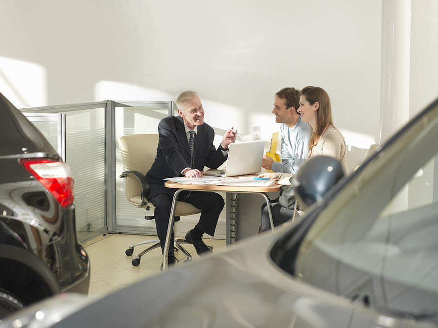 Salesman with laptop in car showroom talking with male and female customers Photograph by Monty Rakusen