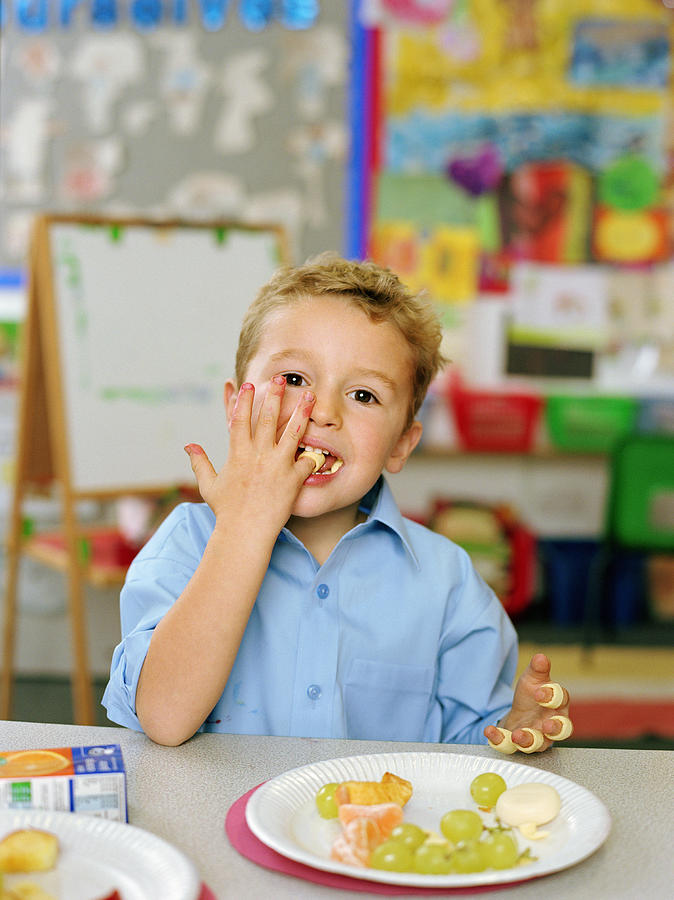 Schoolboy (4-6) eating at table, wearing crisps on fingers, portrait Photograph by Katy McDonnell