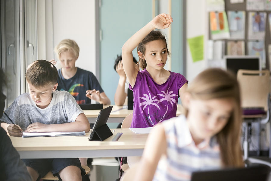 Schoolgirl raising hand at desk in classroom Photograph by Maskot