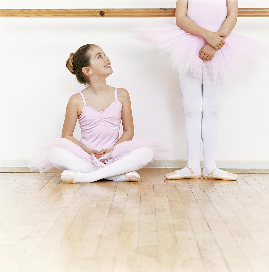 Seated Young Ballet Dancer Looking Up at Another Ballet Dancer Standing in a Dance Studio Photograph by Digital Vision.