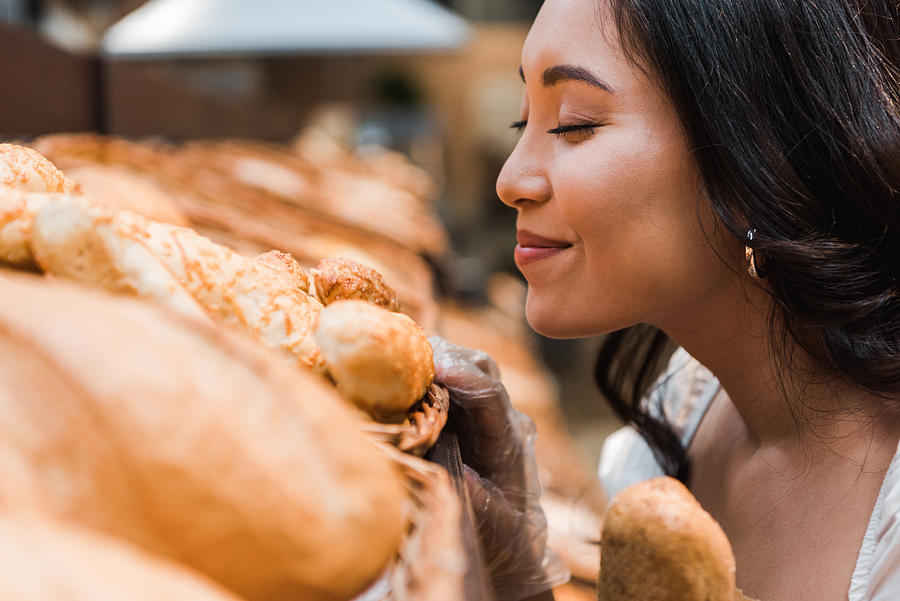 Selective Focus Of Cheerful Asian Woman Smiling While Smelling Bread In Supermarket Photograph by LightFieldStudios