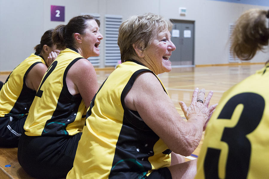 Senior female basketball players sit on bench celebrating beside court Photograph by Jessie Casson