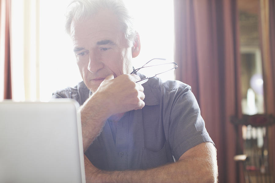 Senior man holding eyeglasses and using laptop Photograph by Sam Edwards