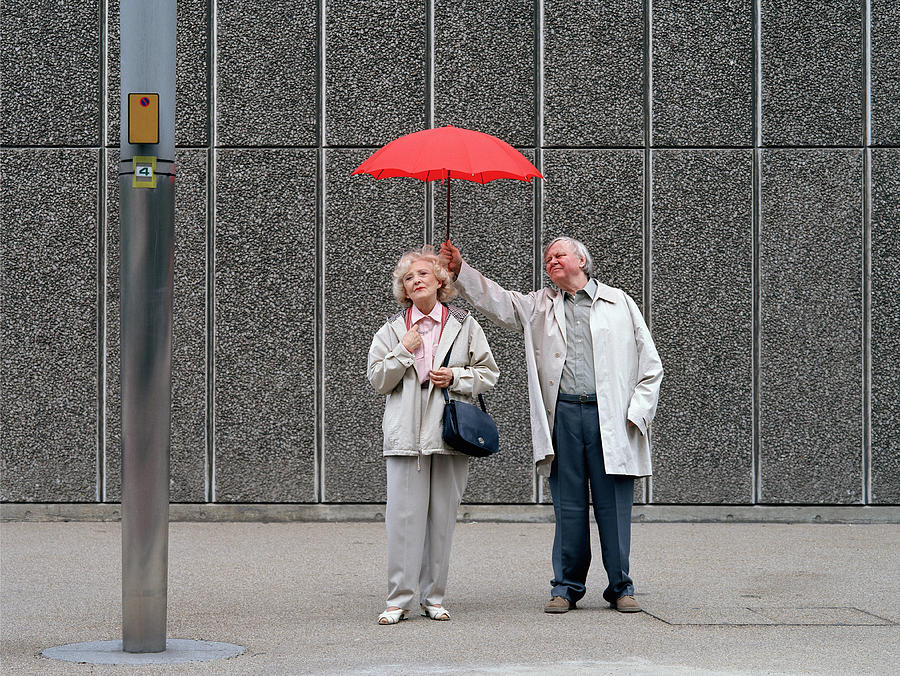 Senior man holding red umbrella over woman, standing on pavement Photograph by Kelvin Murray
