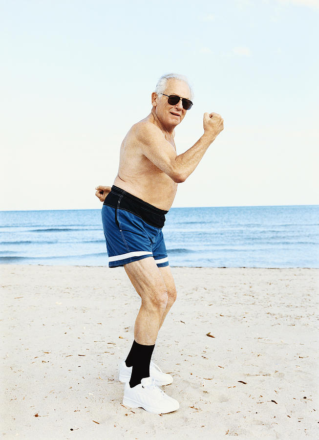 Senior Man in Swimming Trunks Stands on the Beach Flexing His Muscles Photograph by Digital Vision.