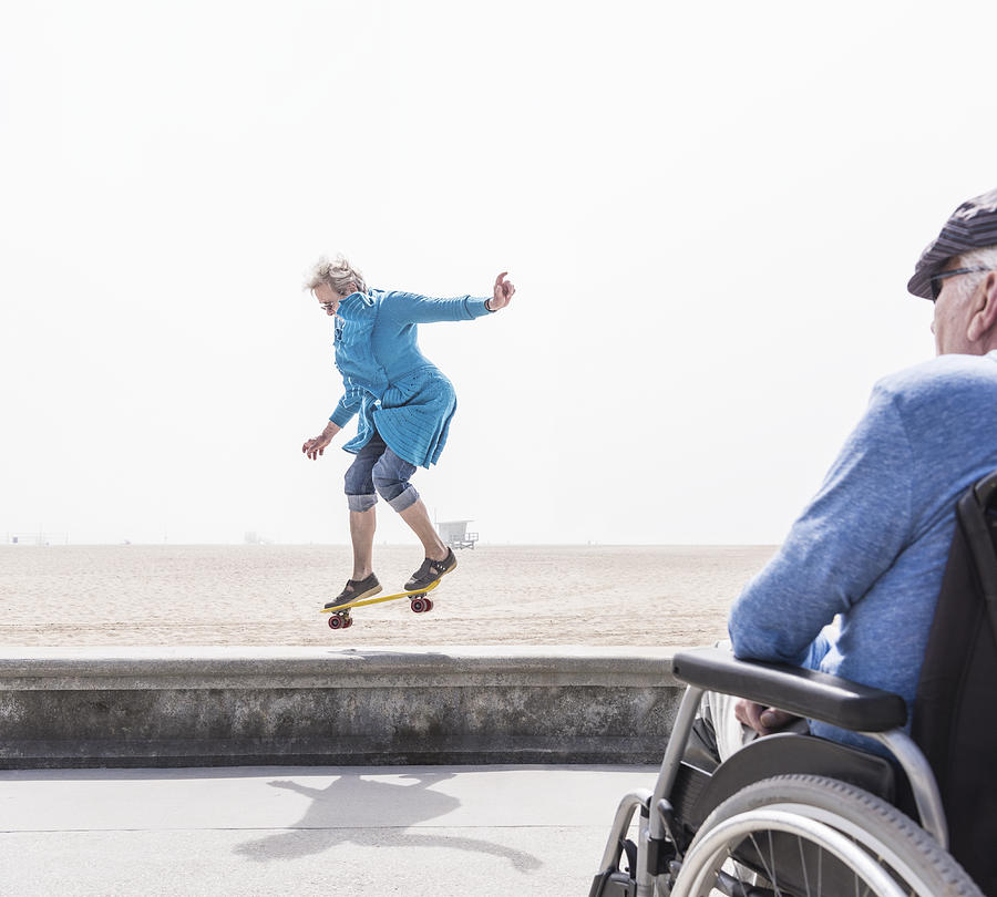 Senior man in wheelchair watching wife doing skateboard trick at beach, Santa Monica, California, USA Photograph by Jlph