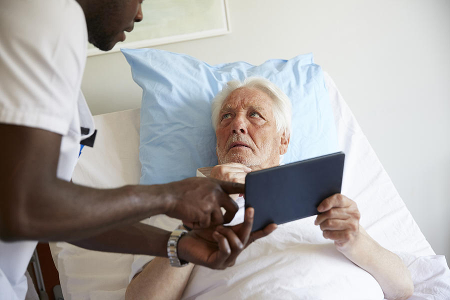 Senior man looking at male nurse while using digital tablet on bed in hospital ward Photograph by Maskot