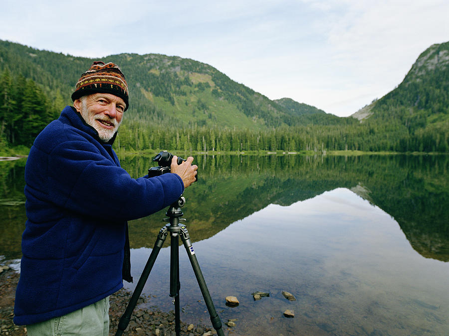 Senior man setting up camera by lake, looking over shoulder Photograph by Mike Powell