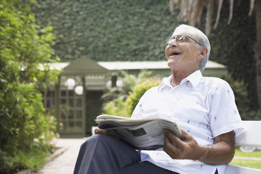 Senior man sitting outdoors with newspaper laughing Photograph by Sam Edwards
