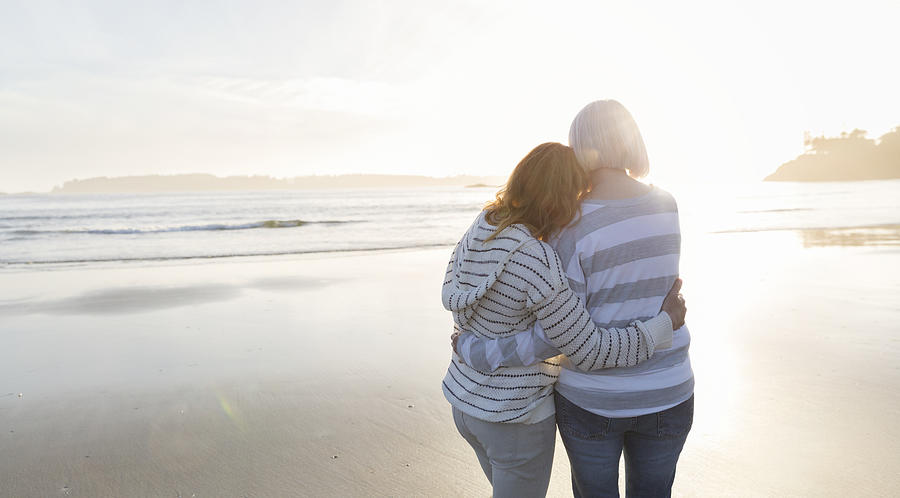 Senior woman and daughter hugging on beach looking at ocean view at sunset Photograph by Compassionate Eye Foundation/Steven Errico