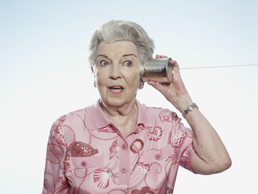 Senior woman comically strains to listen as she holds a tin can phone to her head Photograph by Carl Smith