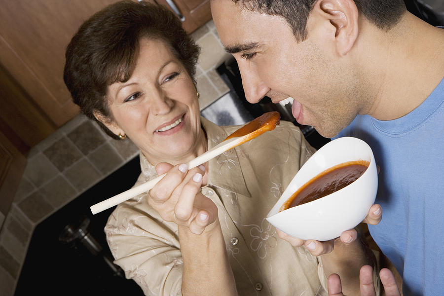 Senior woman feeding tomato soup to her son in the kitchen Photograph by Glowimages