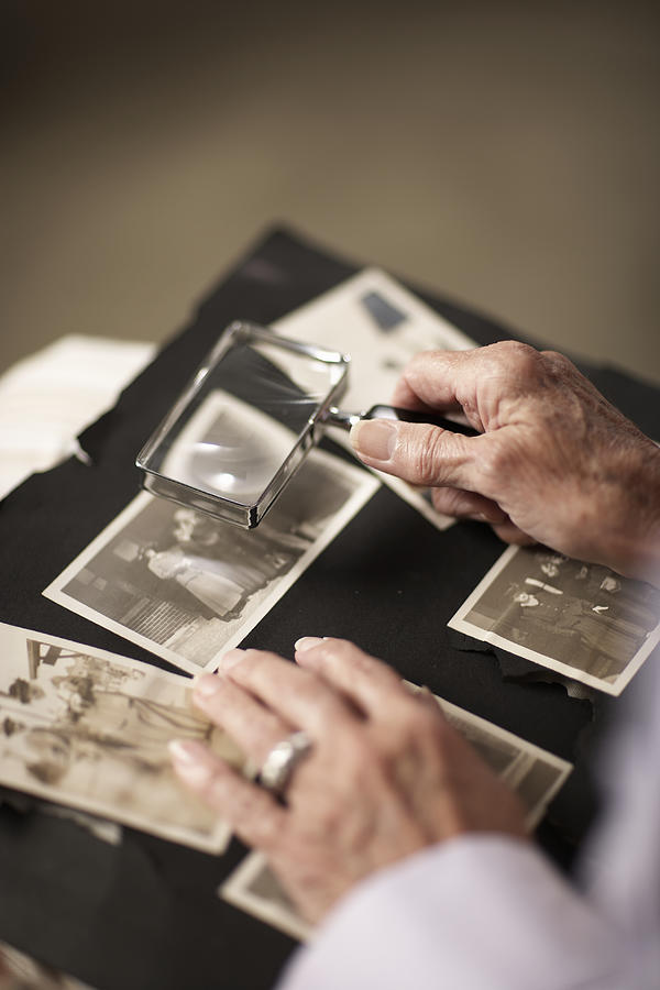 Senior woman looking at old photographs in album through magnifying glass, close-up of hands Photograph by Thomas Northcut