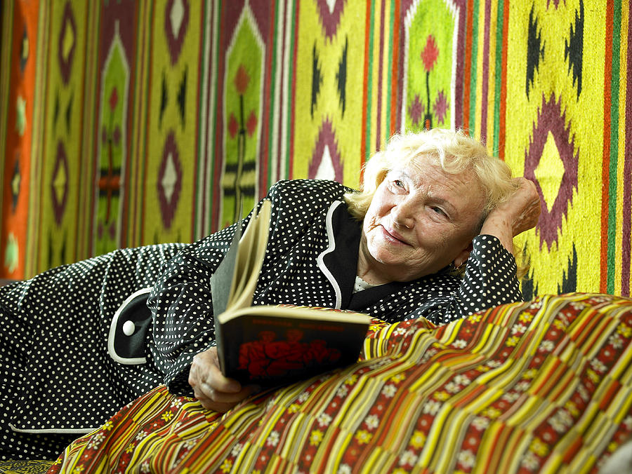Senior woman reclining on bed holding book, smiling Photograph by Hans Neleman